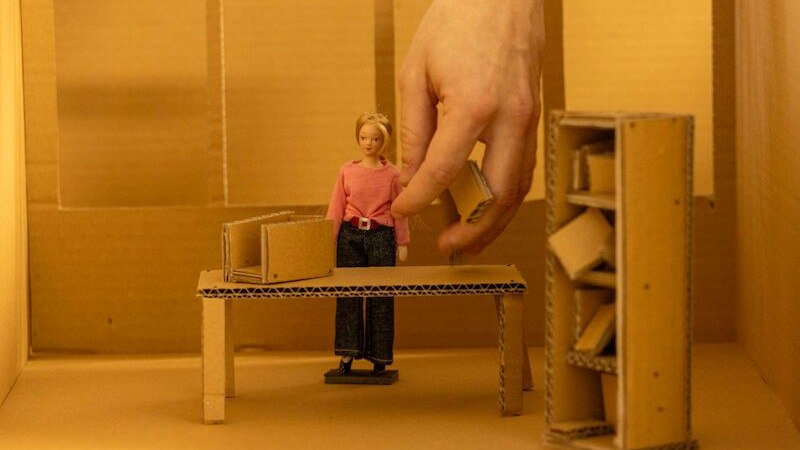 Eine Hand arrangiert eine Puppenfigur in einem Miniaturb&uuml;ro aus Pappe. Die Szene zeigt einen Tisch und ein Regal, alles aus Karton gefertigt., &copy; Theater Rampe