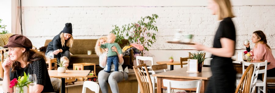 Gem&uuml;tliches Caf&eacute; mit spielenden Kindern und Erwachsenen. Eine Bedienung serviert Getr&auml;nke. Pflanzen und Holzm&ouml;bel schaffen eine einladende Atmosph&auml;re., &copy; Sven Weber