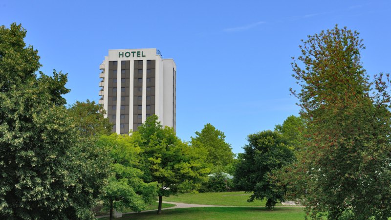 Hotelgebäude hinter Bäumen in einem Park, blauer Himmel., © AMBER HOTEL Leonberg / Stuttgart