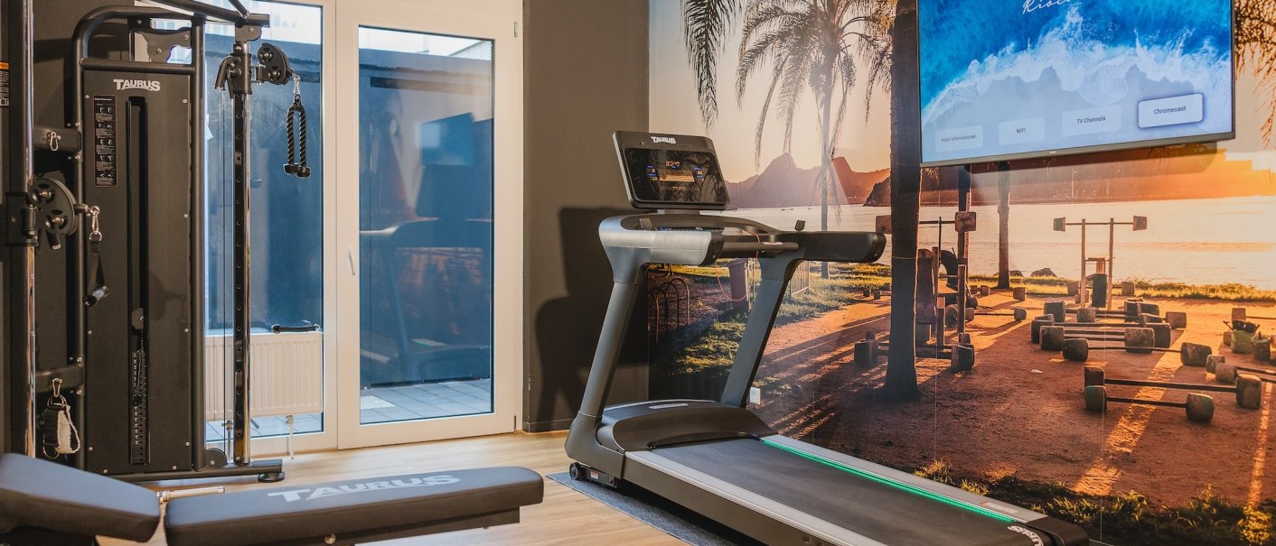 Fitnessraum mit Laufband, Kraftstation und Wandbild eines Strandes bei Sonnenuntergang. Ein Bildschirm zeigt ein Menü an. Heller Holzboden., © TOMAS
