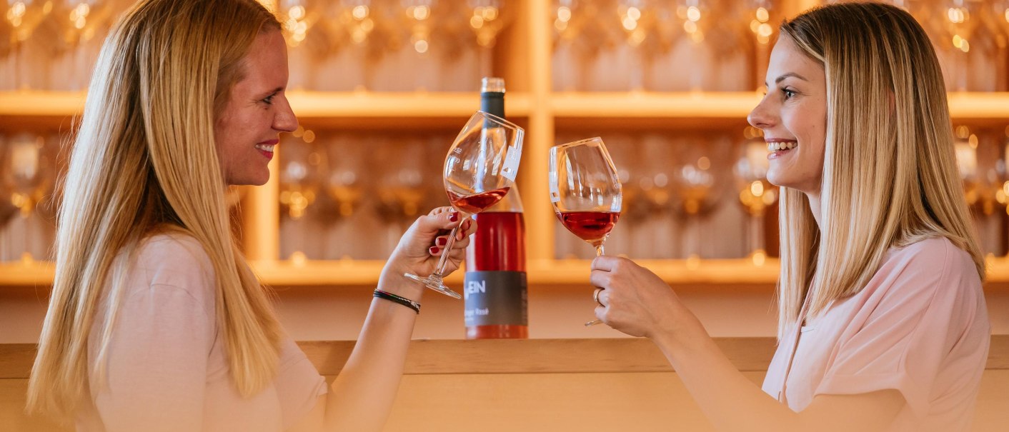 Two women clink glasses of wine in a wine shop. Wine bottles and glasses can be seen in the background., © Stuttgart-Marketing GmbH, Thomas Niedermüller Two women clink glasses of wine in a wine shop. Wine bottles and glasses can be seen in the background., © Stuttgart-Marketing GmbH, Thomas Niedermüller