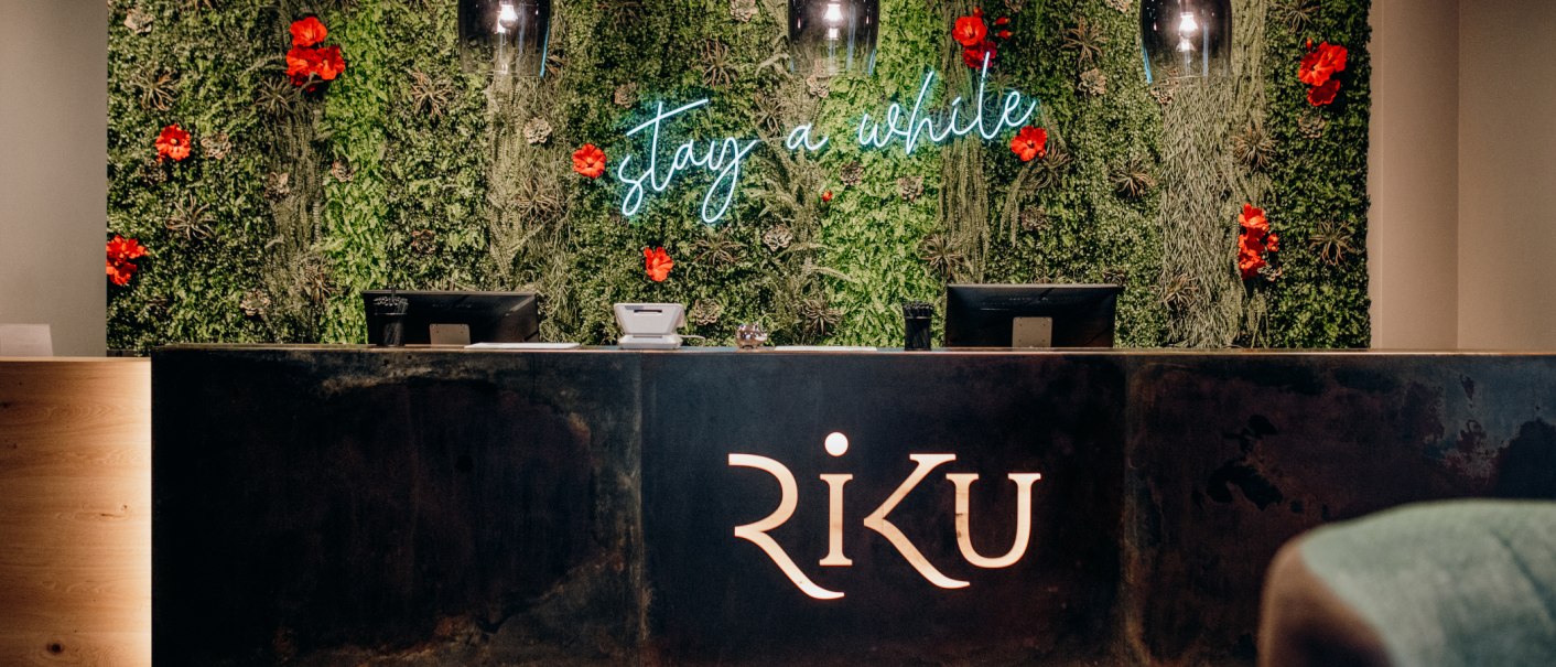 Rezeption mit gr&uuml;nem Pflanzenhintergrund, roten Blumen und Leuchtschrift 'stay a while'. Der Tresen tr&auml;gt das Logo 'RiKU'., &copy; 13 6 Design GmbH (Judenhof 10, 89073 Ulm)