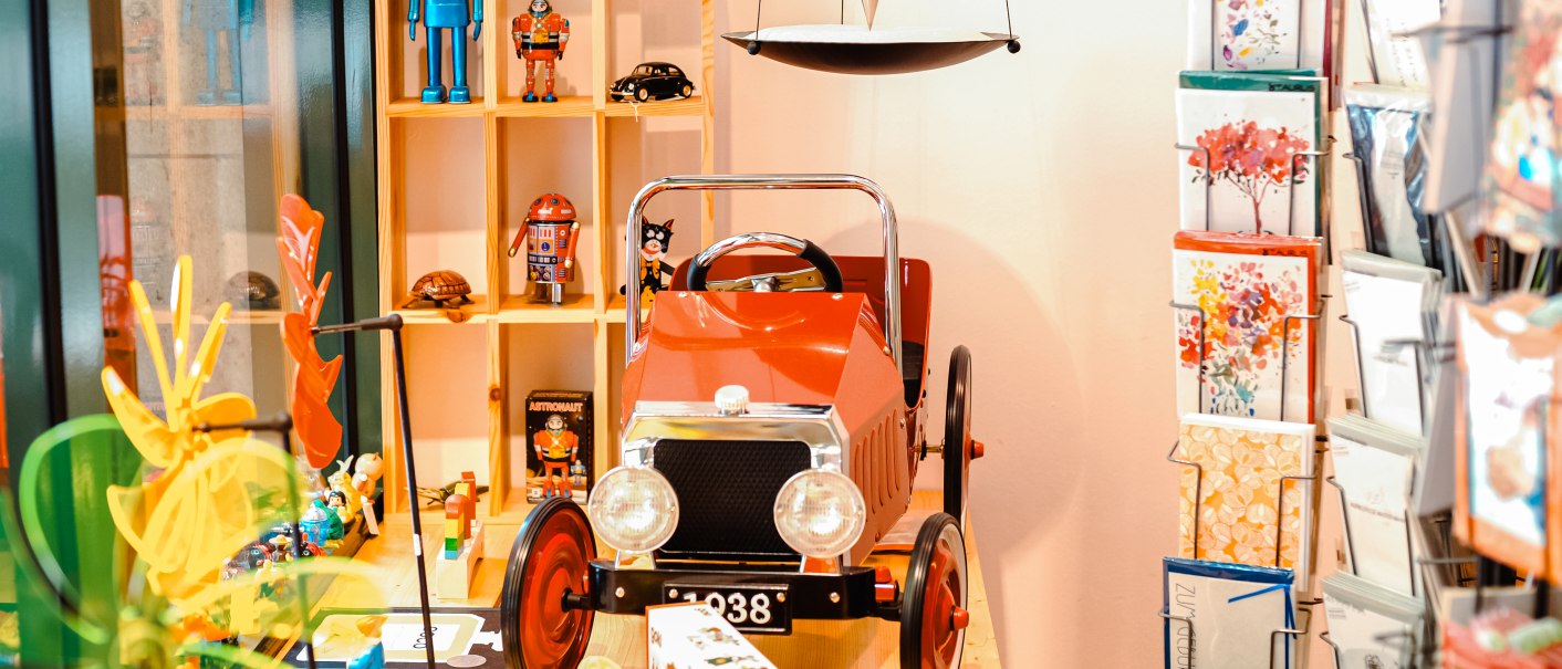 Ein rotes Spielzeugauto steht vor einem Regal mit bunten Figuren. Daneben ein Kartenst&auml;nder mit Gru&szlig;karten., &copy; 1000sch&ouml;n