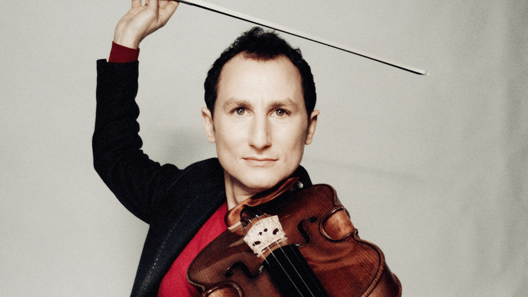 A man holds a violin and raises the bow above his head. He is wearing a dark suit and a red shirt., &copy; Julien Mignot