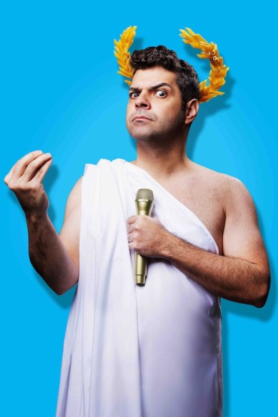 Man in white toga with golden laurel crown and microphone, against a blue background. He gestures with his hand., &copy; Theaterhaus Stuttgart e.V.