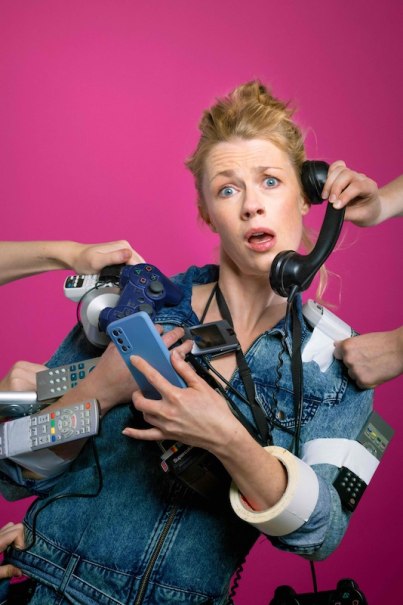 Frau in Jeansjacke h&auml;lt mehrere Telefone, Fernbedienungen und ein Gamepad. Sie wirkt gestresst. Hintergrund ist pink., &copy; Rosenau Kultur e.V.