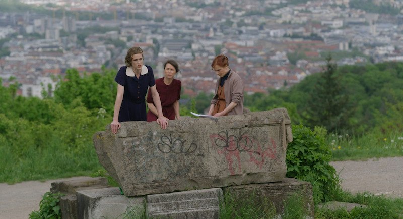 Drei Personen stehen an einem steinernen Denkmal mit Graffiti, im Hintergrund eine Stadtansicht mit vielen Gebäuden und grüner Vegetation., © tri-bühne e.V. Drei Personen stehen an einem steinernen Denkmal mit Graffiti, im Hintergrund eine Stadtansicht mit vielen Gebäuden und grüner Vegetation., © tri-bühne e.V.