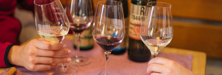 Two people are holding glasses of red and white wine, clinking glasses. In the background are bottles of wine on a table with a purple tablecloth., &copy; 70469R! Weinbau Fabian Rajtschan