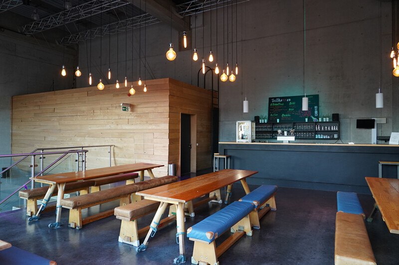 Ein modernes Foyer mit Holztischen und Bänken, einer Bar im Hintergrund und hängenden Glühbirnen als Beleuchtung., © Württembergische Staatstheater Stuttgart Ein modernes Foyer mit Holztischen und Bänken, einer Bar im Hintergrund und hängenden Glühbirnen als Beleuchtung., © Württembergische Staatstheater Stuttgart
