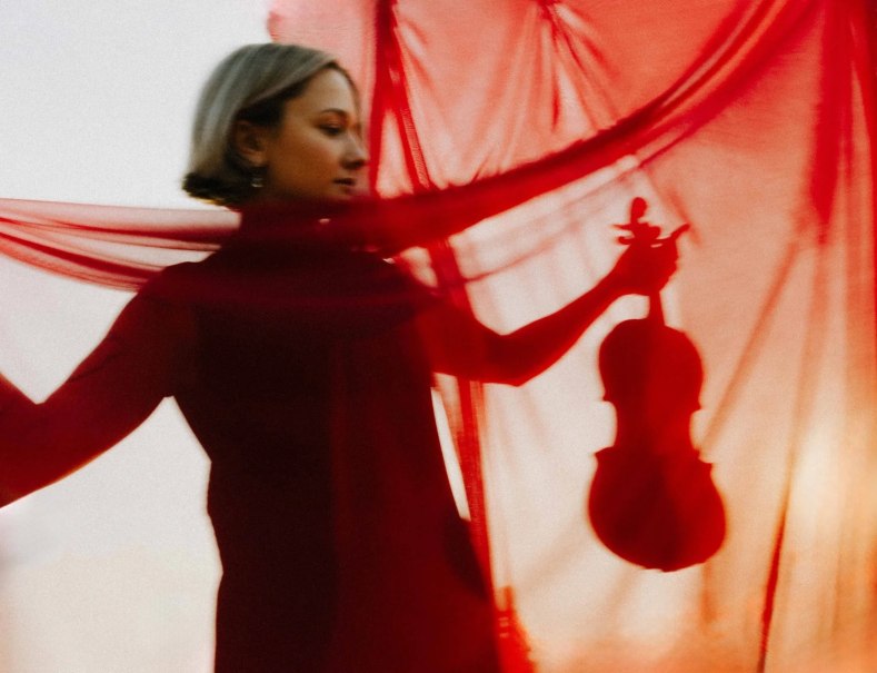 Eine Frau steht hinter einem roten, durchscheinenden Tuch und h&auml;lt eine Violine. Die Szene wirkt k&uuml;nstlerisch und geheimnisvoll., &copy; Sophia Hegewald