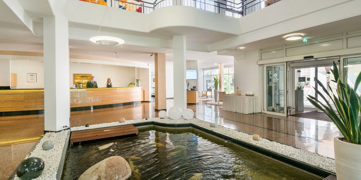 Helle Empfangshalle eines Hotels mit Rezeption, Wasserbecken und Pflanzen. Zwei Personen stehen hinter dem Empfangstresen., © Best Western Plus Hotel am Schlossberg
