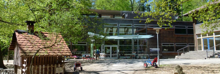Inner courtyard of the House of the Forest with toy vehicles, trees and a small wooden house. Modern glass façade in the background., © Haus des Waldes