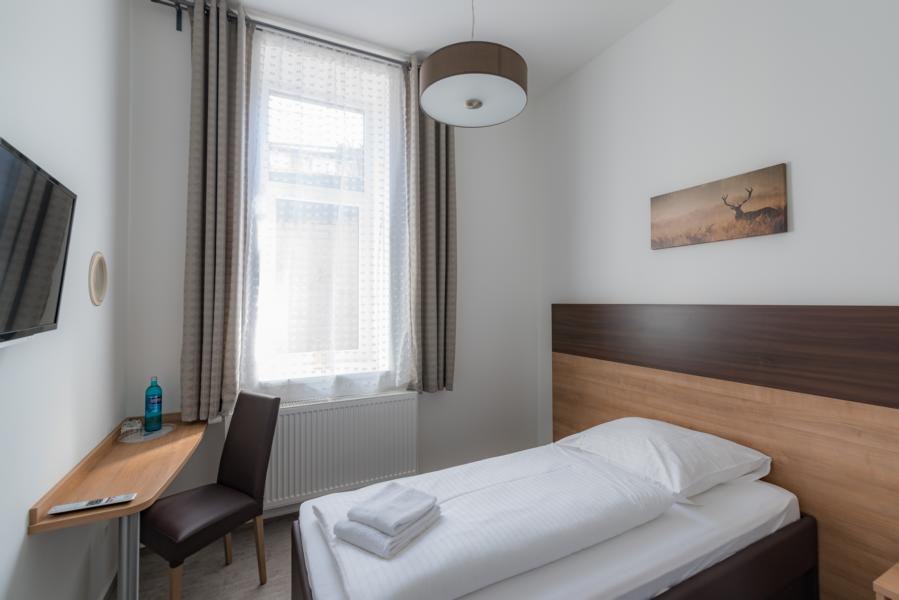 Ein schlichtes Einzelzimmer mit einem Bett, einem kleinen Schreibtisch, einem Stuhl und einem Fenster mit Vorhängen. An der Wand hängt ein Bild., © TOMAS Ein schlichtes Einzelzimmer mit einem Bett, einem kleinen Schreibtisch, einem Stuhl und einem Fenster mit Vorhängen. An der Wand hängt ein Bild., © TOMAS