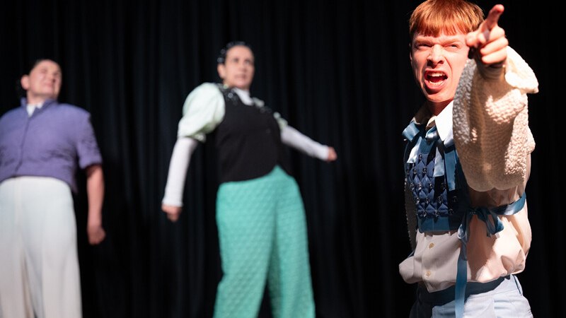 Drei Personen auf einer B&uuml;hne, eine im Vordergrund zeigt energisch nach vorne. Die anderen stehen im Hintergrund vor einem schwarzen Vorhang., &copy; W&uuml;rttembergische Staatstheater Stuttgart