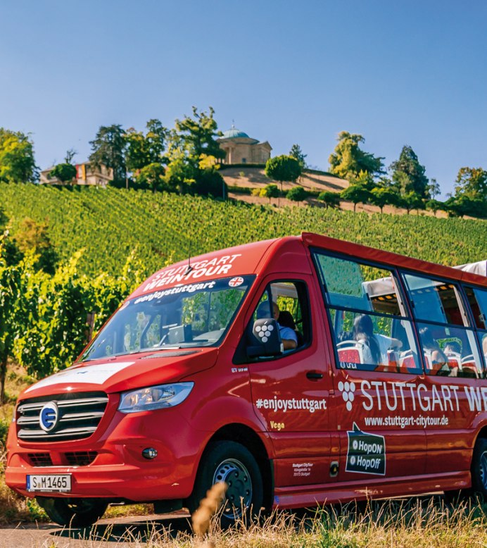 Eine Luftaufnahme zeigt den roten Weintour-Bus der Stuttgarter Citytour, wie er auf einer asphaltierten Straße durch einen Weinberg fährt. Das Laub des Weins ist saftig grün und die Szene ist lichtdurchflutet. , © SMG, Thomas Niedermüller 