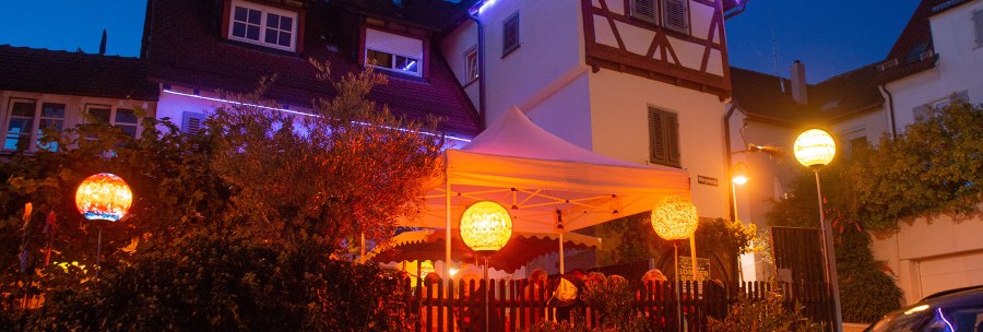 An illuminated half-timbered house with a white tent in the garden. Colorful lanterns and fairy lights create a cozy atmosphere at night., © Weingut Ruoff