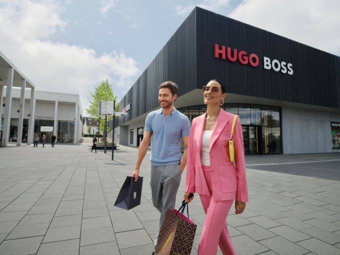 Zwei Personen mit Einkaufstaschen gehen lächelnd vor einem Hugo Boss Outlet in Metzingen. Der Himmel ist leicht bewölkt., © Schlienz-Tours GmbH & Co.KG Zwei Personen mit Einkaufstaschen gehen lächelnd vor einem Hugo Boss Outlet in Metzingen. Der Himmel ist leicht bewölkt., © Schlienz-Tours GmbH & Co.KG