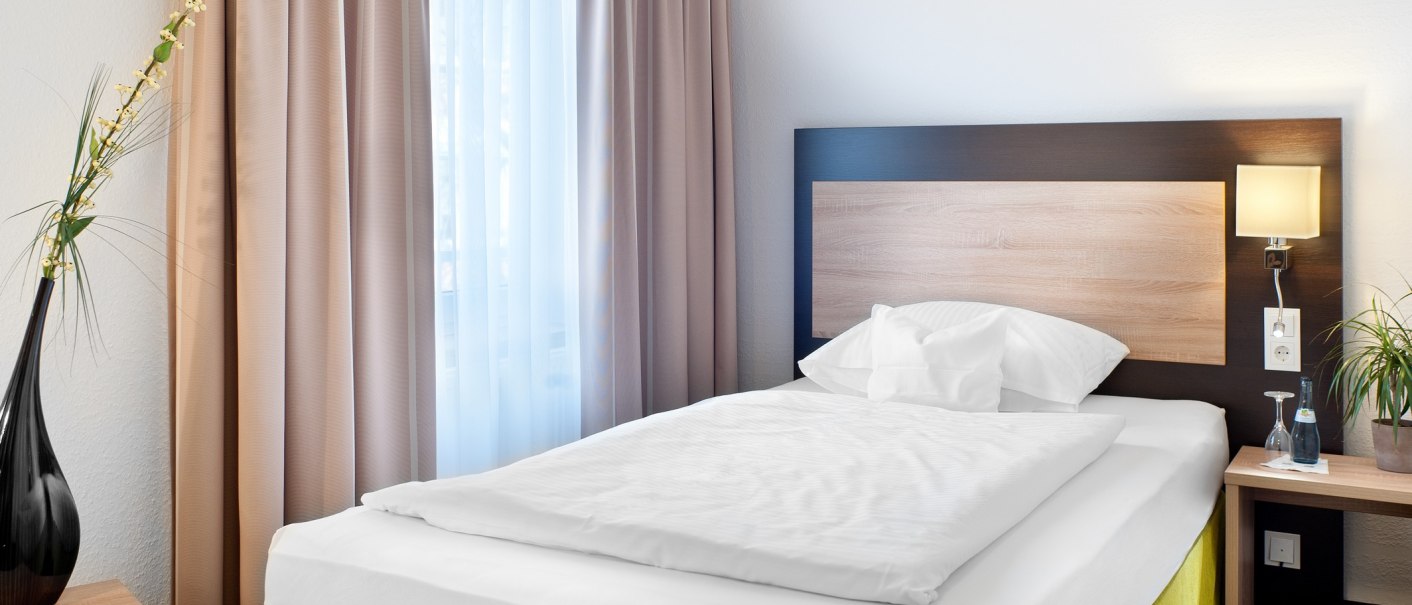 A cozy single room with a single bed, a light-colored wooden headboard, a nightstand, a lamp, and a window with curtains. A vase of flowers sits on a table., &copy; TOMAS