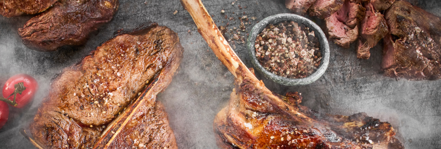 Grilled steaks on a table, surrounded by spices and tomatoes. The steaks are juicy and sprinkled with salt, next to a bowl of peppercorns., © MAREDO