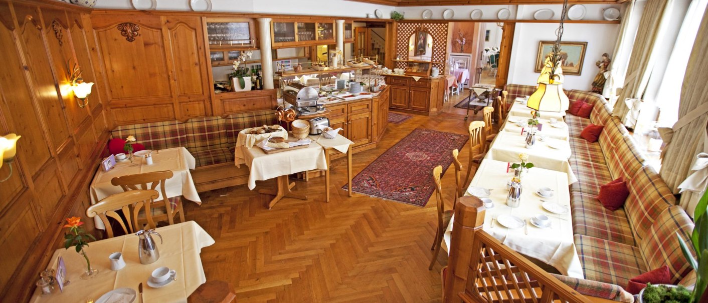 Ein gemütlicher Frühstücksraum mit Holzmöbeln, gedeckten Tischen und karierten Sitzbänken. Ein Buffetbereich ist im Hintergrund zu sehen., © Hotel Hirsch