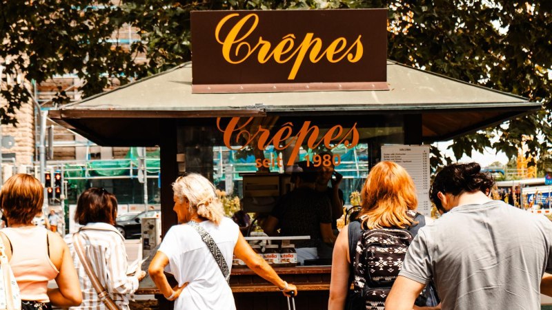 Menschen stehen an einem Crêpes-Stand vor dem Stuttgarter Hauptbahnhof. Der Stand ist mit einem Schild 'Crêpes seit 1980' versehen., © SMG, Sarah Schmid Menschen stehen an einem Crêpes-Stand vor dem Stuttgarter Hauptbahnhof. Der Stand ist mit einem Schild 'Crêpes seit 1980' versehen., © SMG, Sarah Schmid