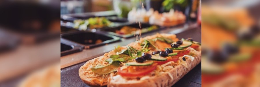 A sandwich is topped with fresh vegetables and olives. A hand sprinkles spices on top. Ingredients in containers can be seen in the background., &copy; FreshSub