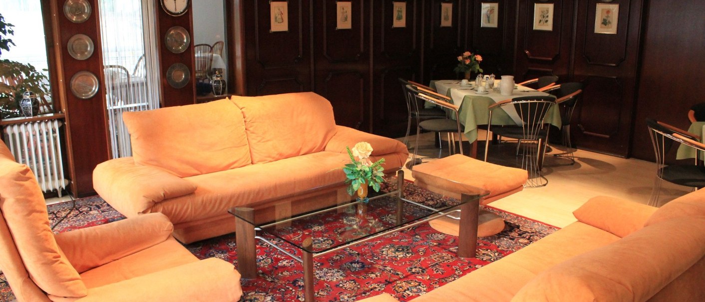 A cozy lounge area in the hotel with orange sofas, a glass table, and a red rug. In the background are set tables and wooden walls with decorations., &copy; Hotel Spahr