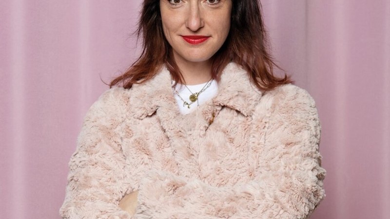 Eine Frau mit braunen Haaren und rotem Lippenstift trägt einen flauschigen, hellen Mantel. Sie steht vor einem rosa Vorhang und lächelt leicht., © Renitenztheater Stuttgart e.V. Eine Frau mit braunen Haaren und rotem Lippenstift trägt einen flauschigen, hellen Mantel. Sie steht vor einem rosa Vorhang und lächelt leicht., © Renitenztheater Stuttgart e.V.