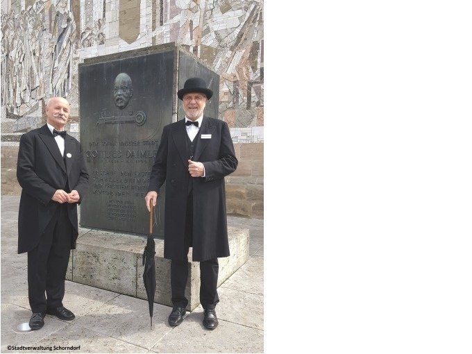 Zwei M&auml;nner in historischer Kleidung posieren vor einem Gottlieb-Daimler-Denkmal in Schorndorf. Sie tragen Anz&uuml;ge und H&uuml;te und halten Regenschirme., &copy; Eigenbetrieb Tourismus und Citymanagement Schorndorf