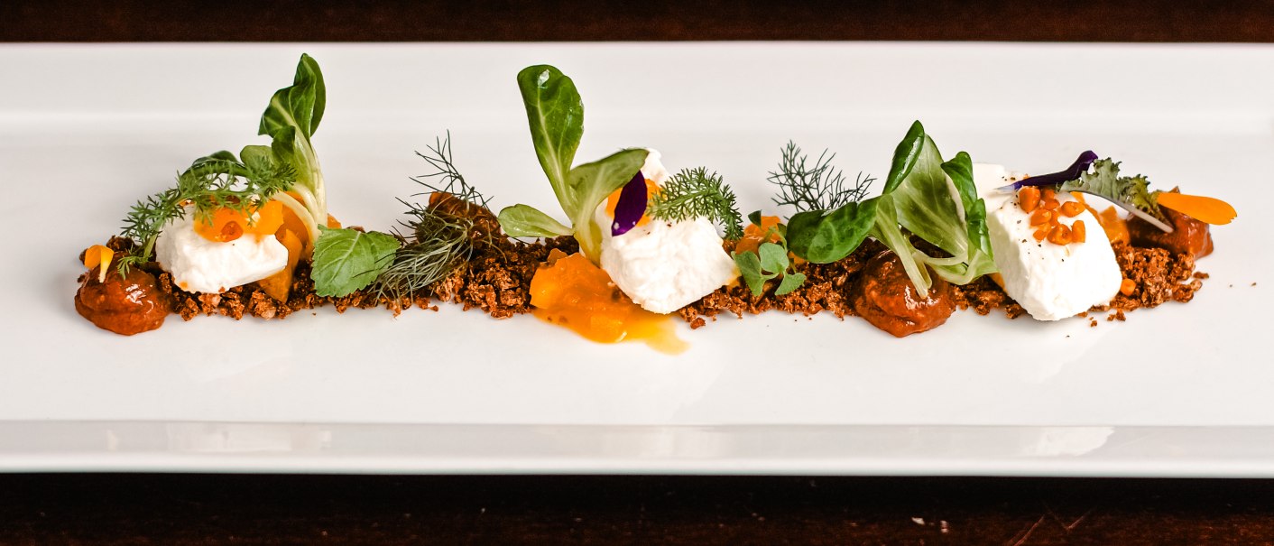 Elegantes Gericht mit Ziegenfrischkäse, Pumpernickel und Feigenchutney, garniert mit frischen Kräutern und Salatblättern auf einem rechteckigen weißen Teller., © Hupperts