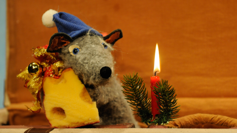 Eine Stoffratte mit blauer Mütze hält ein Stück Käse. Daneben steht eine brennende rote Kerze mit Tannenzweig. Weihnachtliche Szene., © TheaterFusion