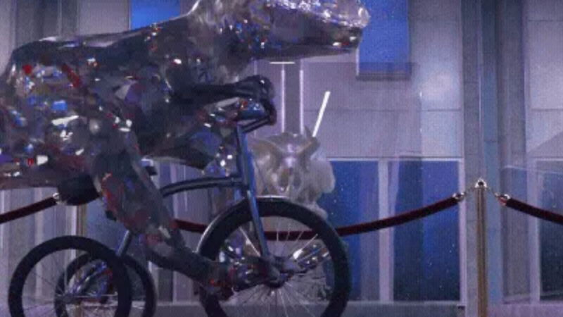 Ein glitzernder Dinosaurier fährt auf einem Fahrrad über einen roten Teppich. Im Hintergrund ist ein modernes Gebäude zu sehen. Logo: KulturRegion Stuttgart., © Adobe Stock, flashmovie