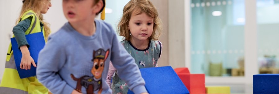 Children play with colorful building blocks on an indoor construction site. They are wearing construction helmets and safety vests. Modern rooms can be seen in the background., &copy; Julia Ochs