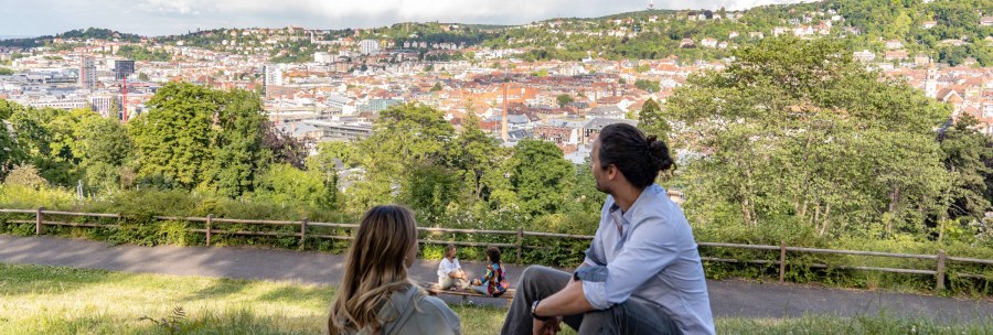 Two people sit on a meadow and look out over the city of Stuttgart. Hills and buildings can be seen in the background, including the television tower., © SMG, Martina Denker Two people sit on a meadow and look out over the city of Stuttgart. Hills and buildings can be seen in the background, including the television tower., © SMG, Martina Denker
