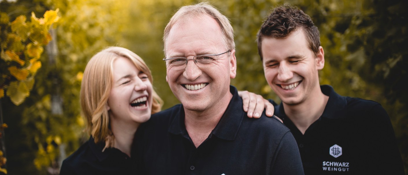 Three people in black clothing are laughing heartily. They are standing in front of a blurred green background. One shirt reads 'Schwarz Weingut'., © Weingut Schwarz