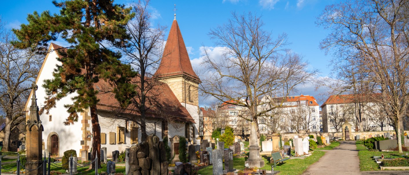 Der Uff-Kirchhof mit Kirche, Grabsteinen und B&auml;umen unter blauem Himmel; im Hintergrund stehen Wohnh&auml;user., &copy; Peter Jagusch/Landeshauptstadt Stuttgart