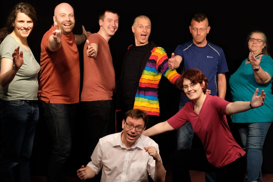 Acht Personen posieren fröhlich vor einem schwarzen Hintergrund. Eine Person trägt eine bunte Jacke., © kaba-reh production Acht Personen posieren fröhlich vor einem schwarzen Hintergrund. Eine Person trägt eine bunte Jacke., © kaba-reh production