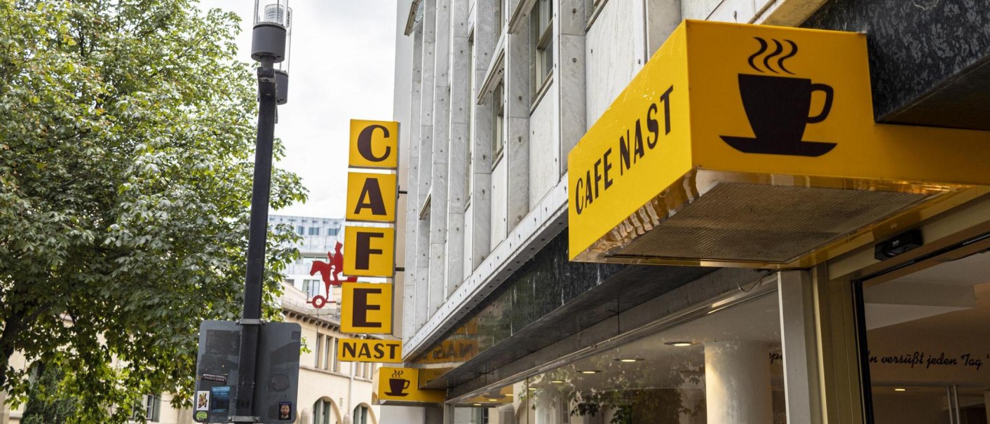 Yellow Cafe Nast sign with coffee cup logo on a building surrounded by trees., © SMG, Sarah Schmid