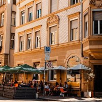 Straßencafé mit Sonnenschirmen vor einem historischen Gebäude. Menschen sitzen draußen, genießen das Wetter. Verkehrsschilder und eine Vespa sind sichtbar., © Stuttgart Marketing GmbH, Sarah Schmid