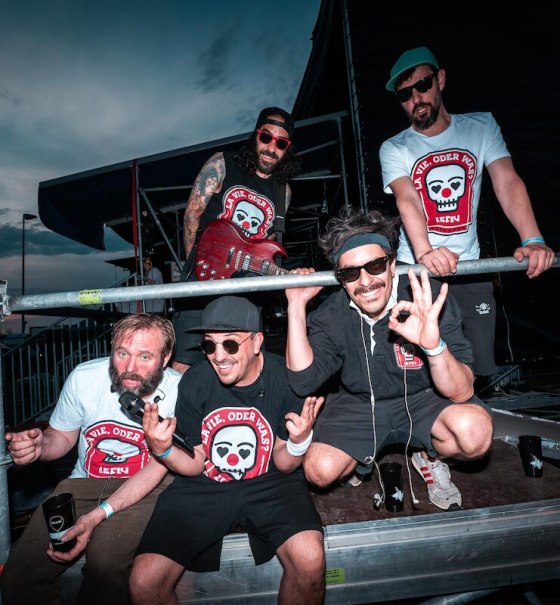 F&uuml;nf M&auml;nner posieren fr&ouml;hlich auf einer B&uuml;hne, tragen T-Shirts mit Totenkopf-Logo und l&auml;cheln in die Kamera. Einer h&auml;lt eine Gitarre., &copy; clubCANN - Stuttgarter Jugendhaus gGmbH