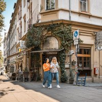 Zwei Frauen spazieren lächelnd am Café Einstein vorbei. Die Straße ist gesäumt von Bäumen und Außensitzplätzen. Es herrscht eine entspannte Atmosphäre., © Stuttgart-Marketing GmbH, Sarah Schmid Zwei Frauen spazieren lächelnd am Café Einstein vorbei. Die Straße ist gesäumt von Bäumen und Außensitzplätzen. Es herrscht eine entspannte Atmosphäre., © Stuttgart-Marketing GmbH, Sarah Schmid
