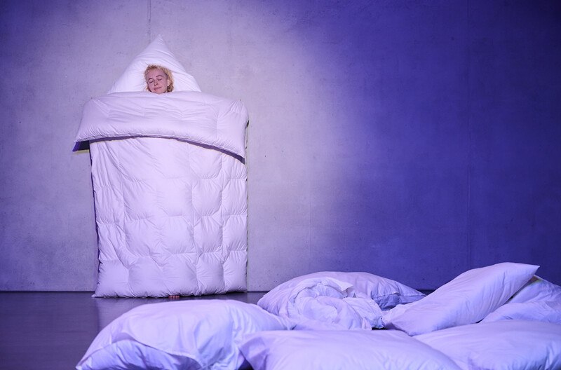 A person stands against a wall wrapped in a blanket, surrounded by cushions on the floor. The scene is bathed in blue light., © Württembergische Staatstheater Stuttgart A person stands against a wall wrapped in a blanket, surrounded by cushions on the floor. The scene is bathed in blue light., © Württembergische Staatstheater Stuttgart