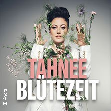 Person in weißem Anzug, umgeben von Blumen, vor grauem Hintergrund. Text: TAHNEE BLÜTEZEIT., © links im Bild