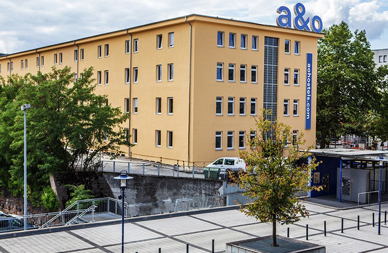 Gelbes Gebäude mit a&o Hostel Logo, umgeben von Bäumen und einem gepflasterten Platz. Im Vordergrund ein Baum und Laternen., © A&O Hostels Marketing GmbH