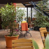 Gem&uuml;tlicher Caf&eacute;-Au&szlig;enbereich mit Holztischen und -st&uuml;hlen, umgeben von Pflanzen. Bunte Decken auf den St&uuml;hlen sorgen f&uuml;r eine einladende Atmosph&auml;re., &copy; SMG, Sarah Schmid