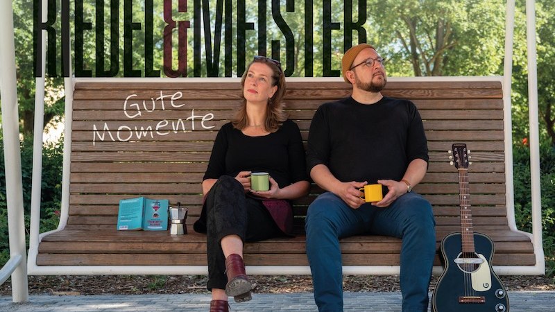 Zwei Personen sitzen auf einer Bank mit Kaffeetassen, eine Gitarre steht daneben. Im Hintergrund der Schriftzug 'Riedel & Meister' und 'Gute Momente'., &copy; Rosenau Kultur e.V.