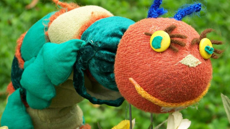 Bunte Stoffraupe mit rotem Kopf, gelben Augen und blauen F&uuml;hlern vor gr&uuml;nem Hintergrund. Sie wirkt fr&ouml;hlich und kindgerecht gestaltet., &copy; Nellys Puppentheater