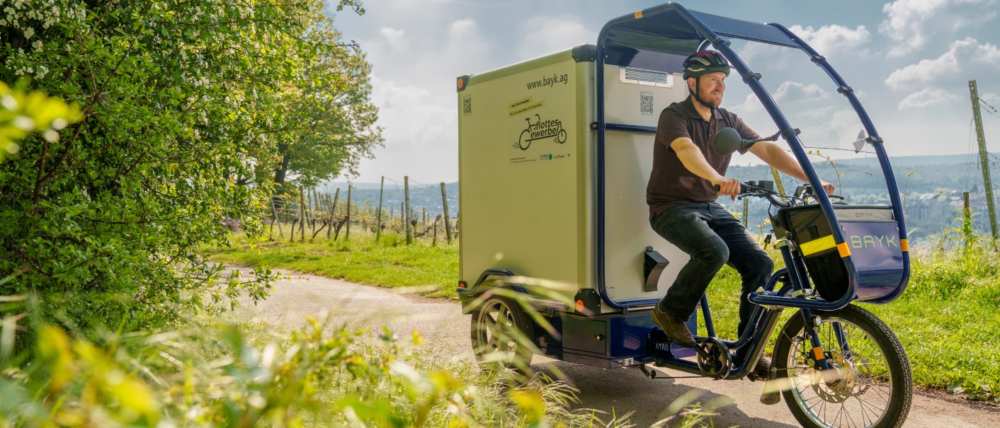 A man rides a cargo bike on a rural road, surrounded by green nature and trees. The sky is slightly cloudy., &copy; 70469R!