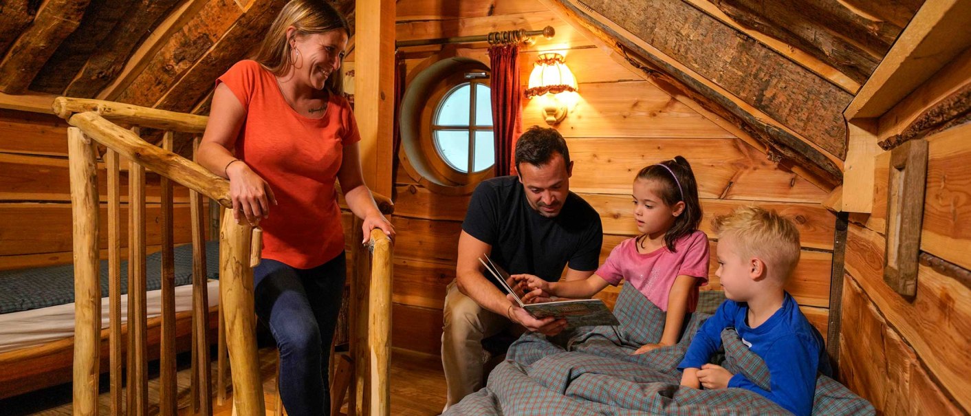 A family in a cozy treehouse. The father is reading to the children while the mother watches with a smile. Wooden walls and rustic furnishings., &copy; Tripsdrill Natur-Resort GmbH