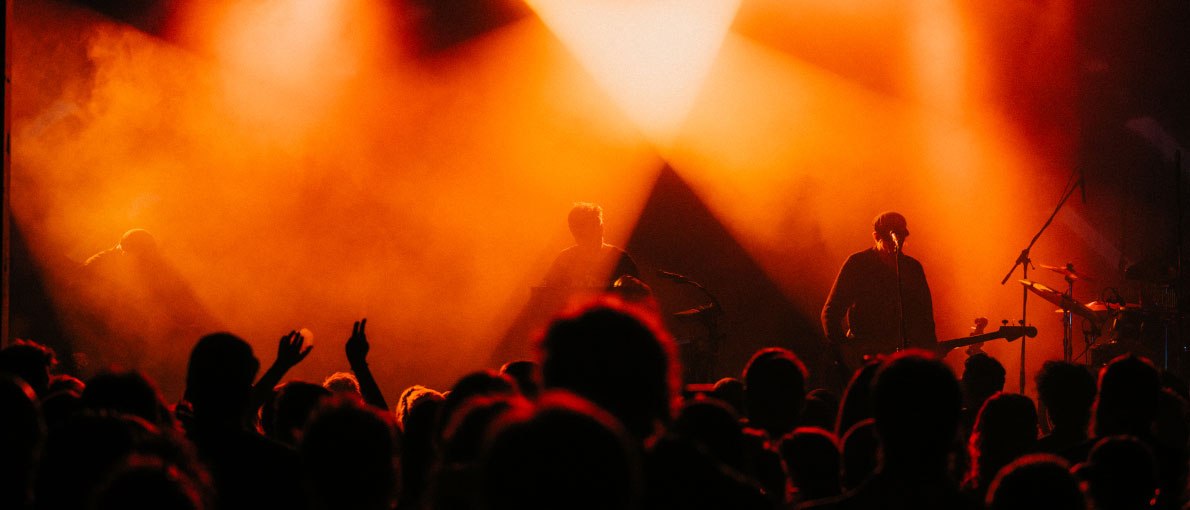 Eine Band spielt auf einer B&uuml;hne, beleuchtet von intensivem orangefarbenem Licht, w&auml;hrend das Publikum im Vordergrund jubelt., &copy; Madlen Medvedovskyy